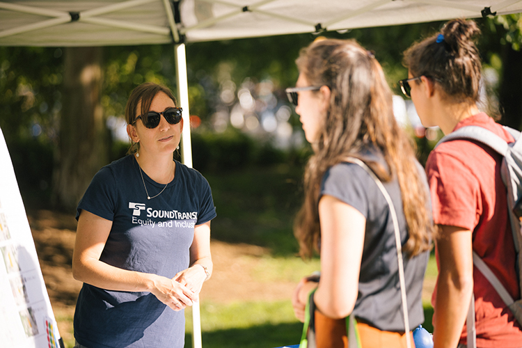 A Sound Transit employee stands and talks to two people visiting the project booth at an outdoor event. 
