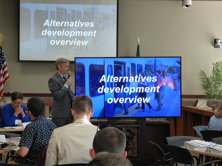 有一人站在两个大屏幕旁，屏幕上方写着 “Alternatives “开发总览”，此人拿着麦克风在一个大房间里对着一群人讲话. 