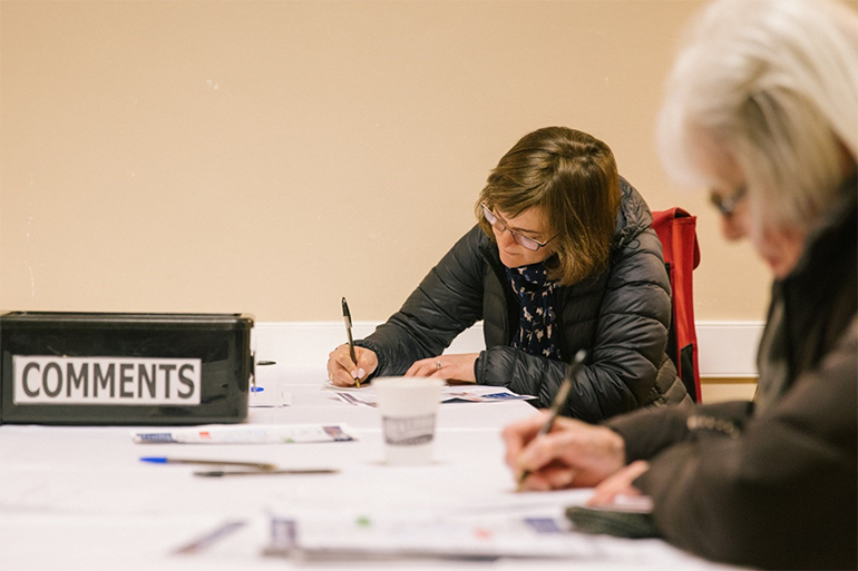 Two people fill out comment submission forms while seated at a table with a box labeled ,comments' nearby.